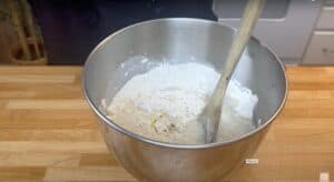 Mixing bread dough ingredients in a metal bowl with wooden spoon on a wooden countertop.